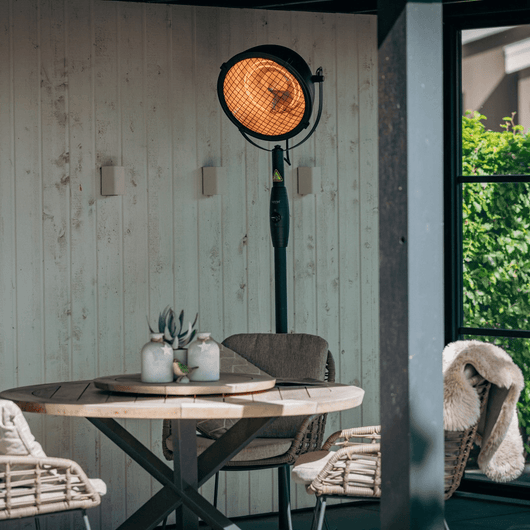 Zwarte staande heater met draaibare kop bij een tuinsetje in een overkapping